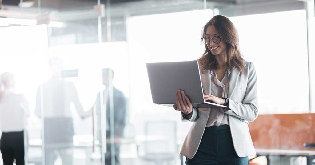 A woman in business attire stands in an office holding a laptop, with AI-powered tools on her screen, while other people and large windows are visible in the background.