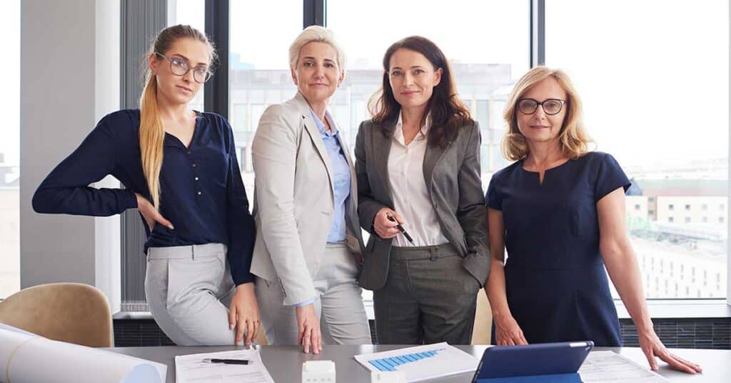 Four women in business attire stand in an office by a table with documents, a tablet, and IPO charts, posing for a group photo in front of large windows.