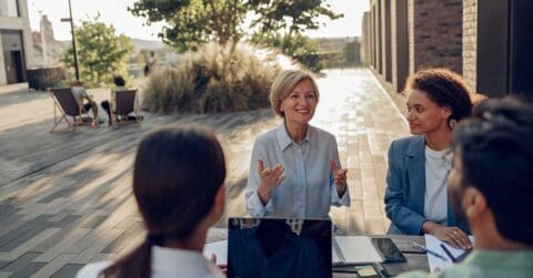 Four people sit at an outdoor table, engaged in a leadership discussion with notebooks and a laptop, surrounded by sunlight and greenery.