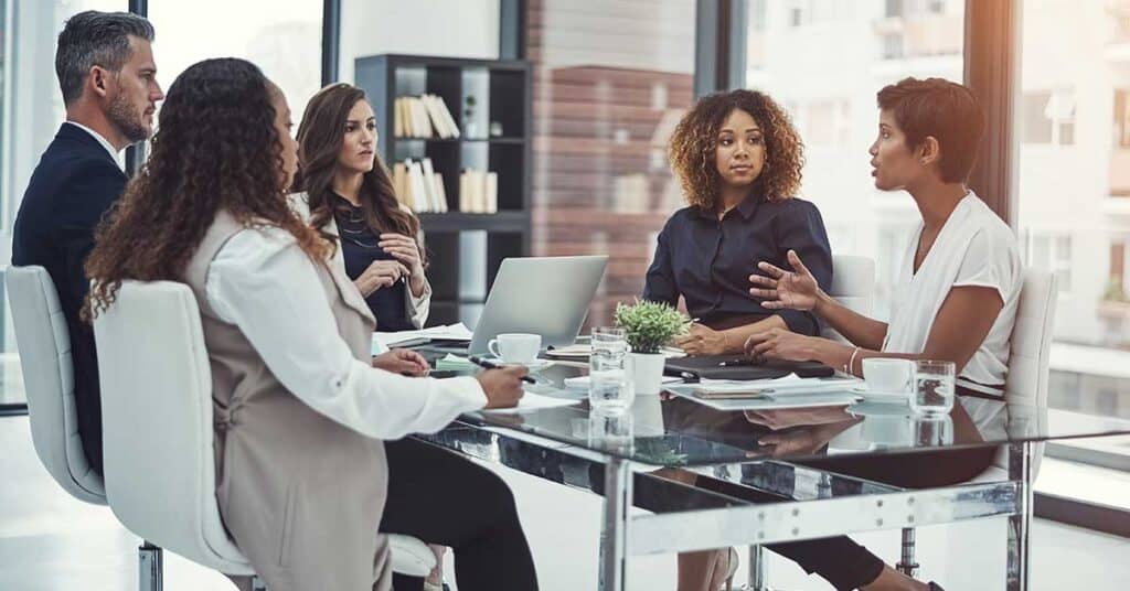 Five people sit around a glass table in a modern office, demonstrating strong leadership as they engage in a business meeting with laptops, documents, and coffee cups present.