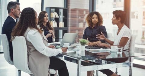 Five people sit around a glass table in a modern office, demonstrating strong leadership as they engage in a business meeting with laptops, documents, and coffee cups present.