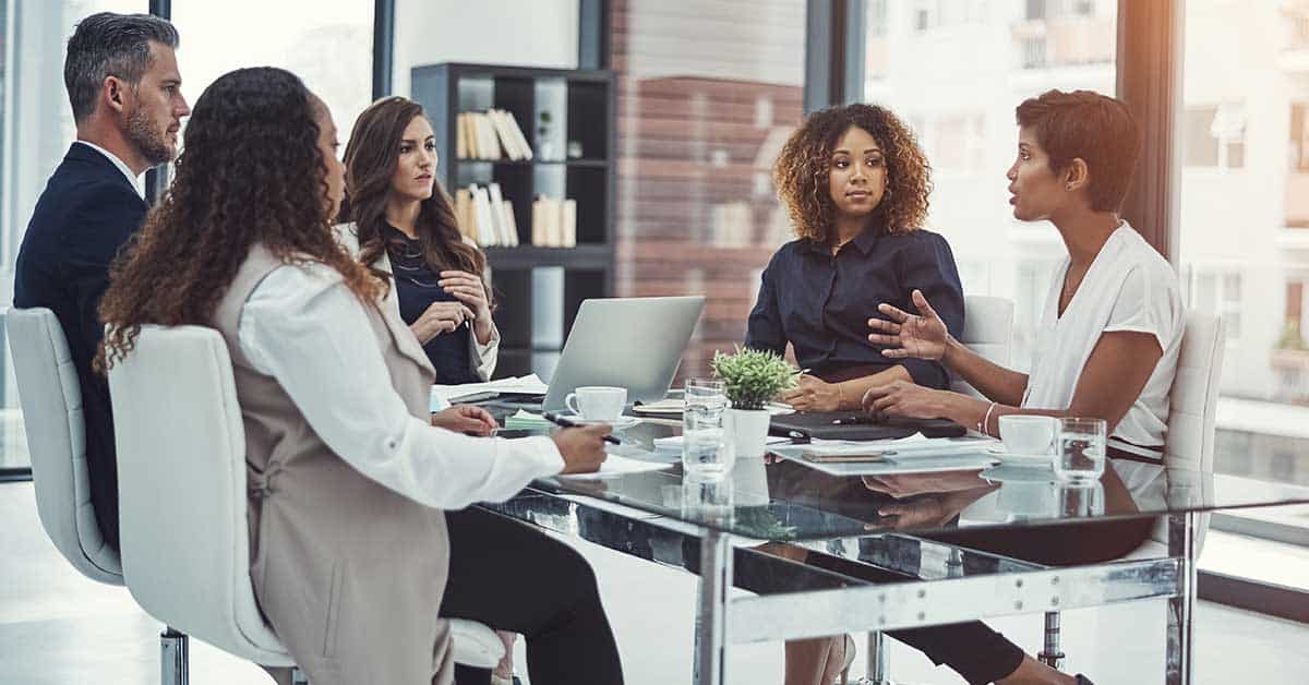 Five people sit around a glass table in a modern office, demonstrating strong leadership as they engage in a business meeting with laptops, documents, and coffee cups present.