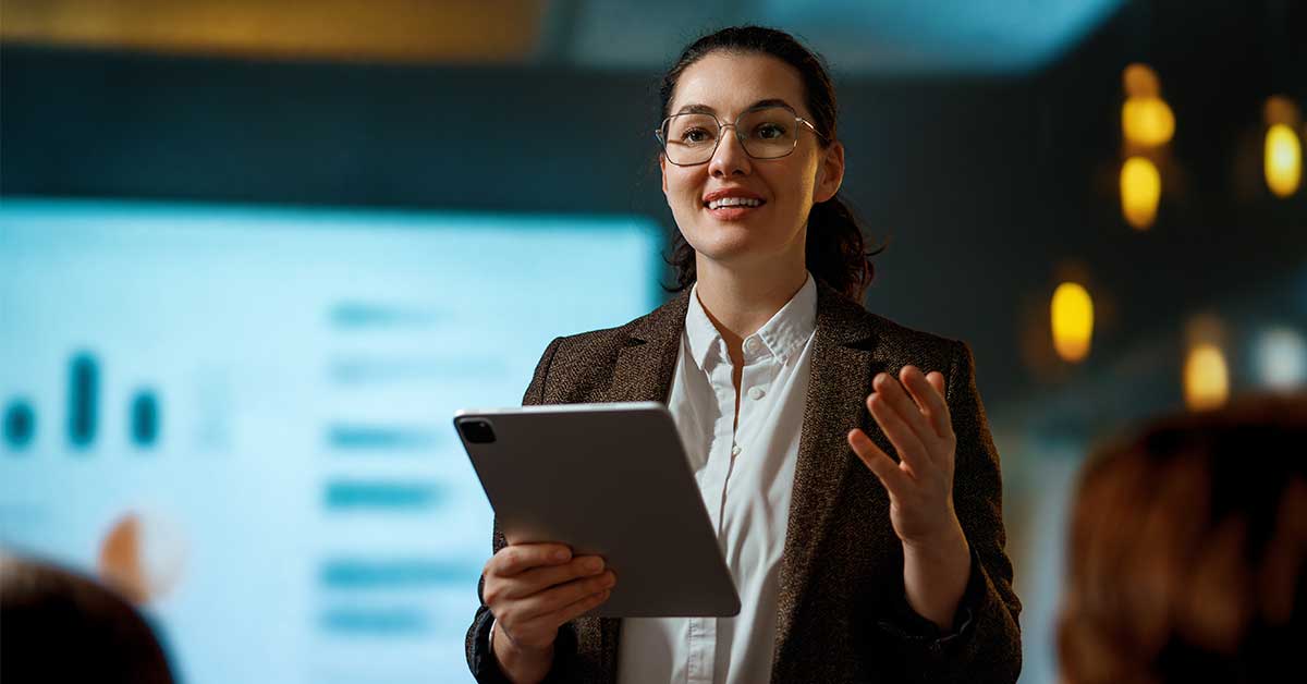 A woman wearing glasses and a blazer holds a tablet and speaks in front of a screen displaying graphs and charts in a dimly lit room.