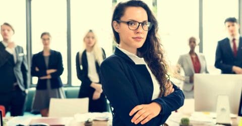 A woman in business attire stands confidently with arms crossed in an office, with a group of colleagues in professional clothing blurred in the background.