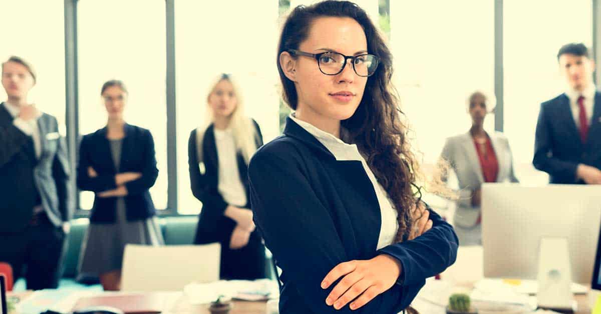 A woman in business attire stands confidently with arms crossed in an office, with a group of colleagues in professional clothing blurred in the background.