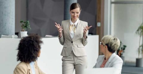 A woman in a light suit demonstrates leadership as she stands and speaks to two seated colleagues in an office setting.