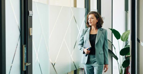 A woman in a light blue suit, possibly discussing sponsorship, holds a notebook while walking down a hallway with frosted glass panels and a potted plant.