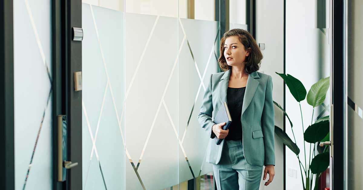 A woman in a light blue suit, possibly discussing sponsorship, holds a notebook while walking down a hallway with frosted glass panels and a potted plant.