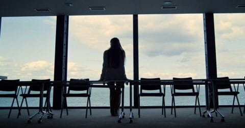 A person stands alone in a conference room, facing large windows with a view of the sea and cloudy sky outside.