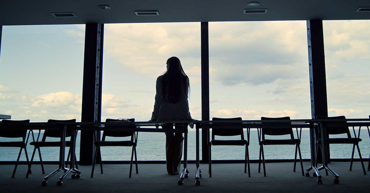 A person stands alone in a conference room, facing large windows with a view of the sea and cloudy sky outside.