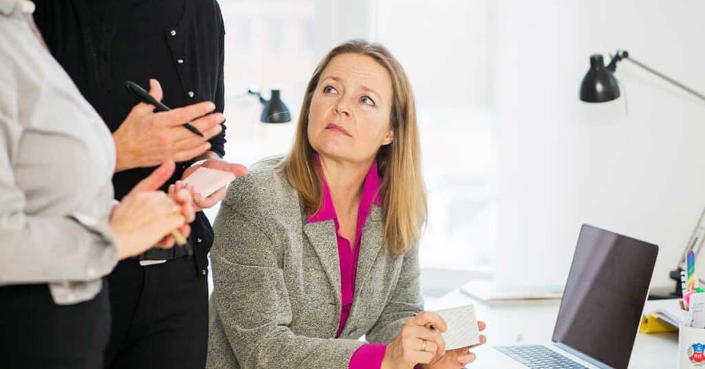 Three people in an office setting discuss something. One woman, likely an executive, sits at a desk with a laptop, listening to two standing colleagues holding notepads and pens.