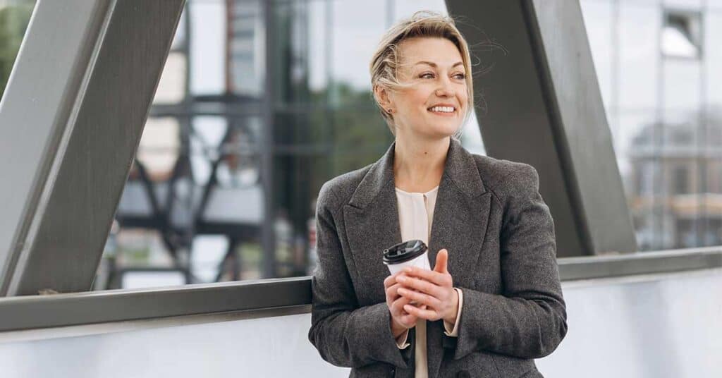 Woman in a gray blazer holding a takeaway coffee cup, standing outdoors near a modern building structure, looking to the side and smiling with confidence and leadership.