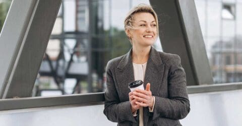 Woman in a gray blazer holding a takeaway coffee cup, standing outdoors near a modern building structure, looking to the side and smiling with confidence and leadership.
