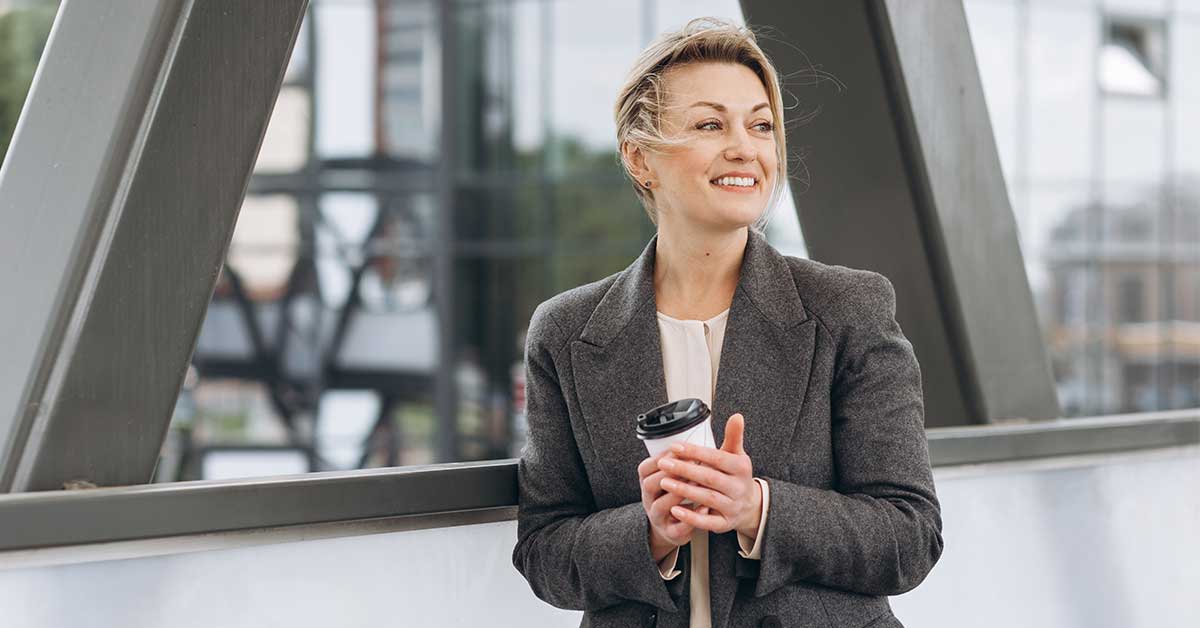 Woman in a gray blazer holding a takeaway coffee cup, standing outdoors near a modern building structure, looking to the side and smiling with confidence and leadership.