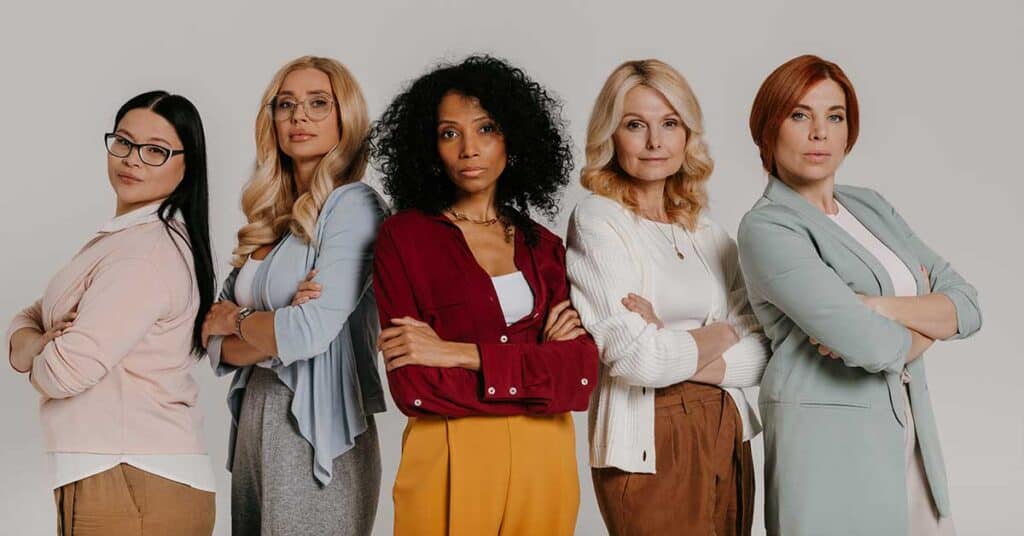 Five women stand side by side with arms crossed, facing the camera against a plain background. Dressed in business casual attire, they convey serious expressions, embodying confidence and leadership in the evolving world of AI.