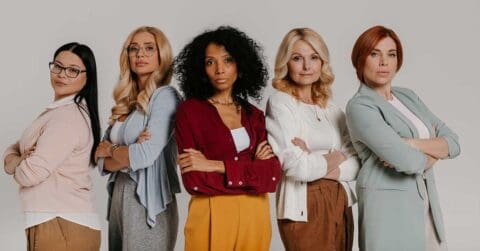 Five women stand side by side with arms crossed, facing the camera against a plain background. Dressed in business casual attire, they convey serious expressions, embodying confidence and leadership in the evolving world of AI.