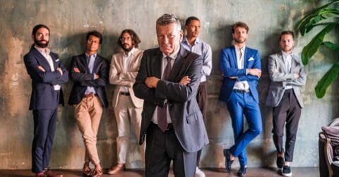 A group of six men in business attire stand against a textured wall, with one man in a suit standing confidently at the front, arms crossed, representing leadership in the age of AI.