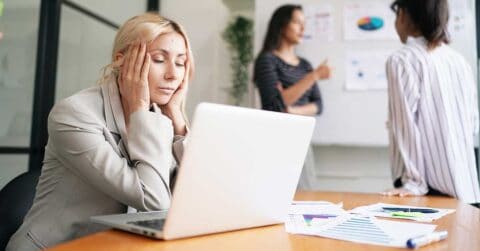 An executive woman sits at a desk with her hands on her face in front of a laptop, while two people discuss charts on a whiteboard in the background. Papers and charts are scattered on the desk.