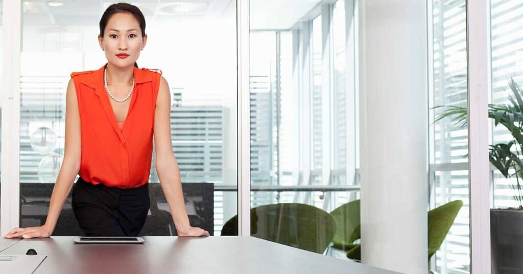 A woman in a red blouse stands confidently at a conference table in a modern office with large windows and minimal decor, exuding professionalism and clear boundaries.