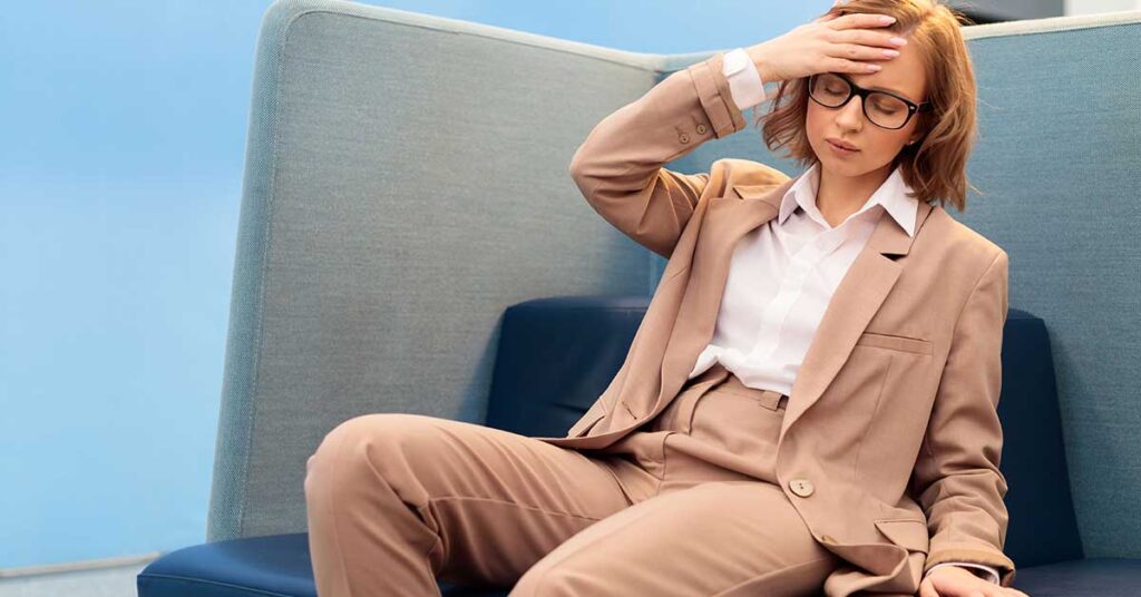 An executive woman in a beige suit and glasses sits on a sofa, touching her forehead with a tired or stressed expression.