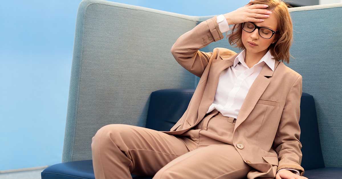 An executive woman in a beige suit and glasses sits on a sofa, touching her forehead with a tired or stressed expression.