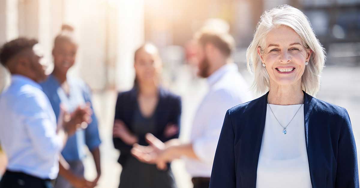 An older woman in business attire smiles at the camera, exuding quiet influence, while a group of four people converse in the blurred background outdoors.