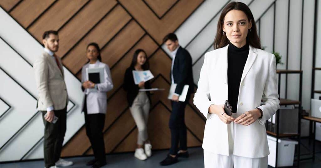 A woman in a white suit exudes leadership as she stands in the foreground, while four colleagues engage in discussion in the background of a modern office.