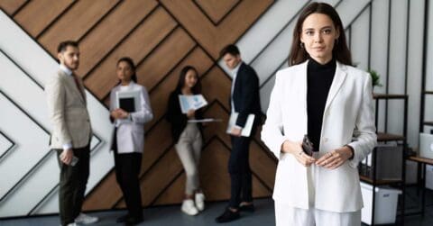A woman in a white suit exudes leadership as she stands in the foreground, while four colleagues engage in discussion in the background of a modern office.