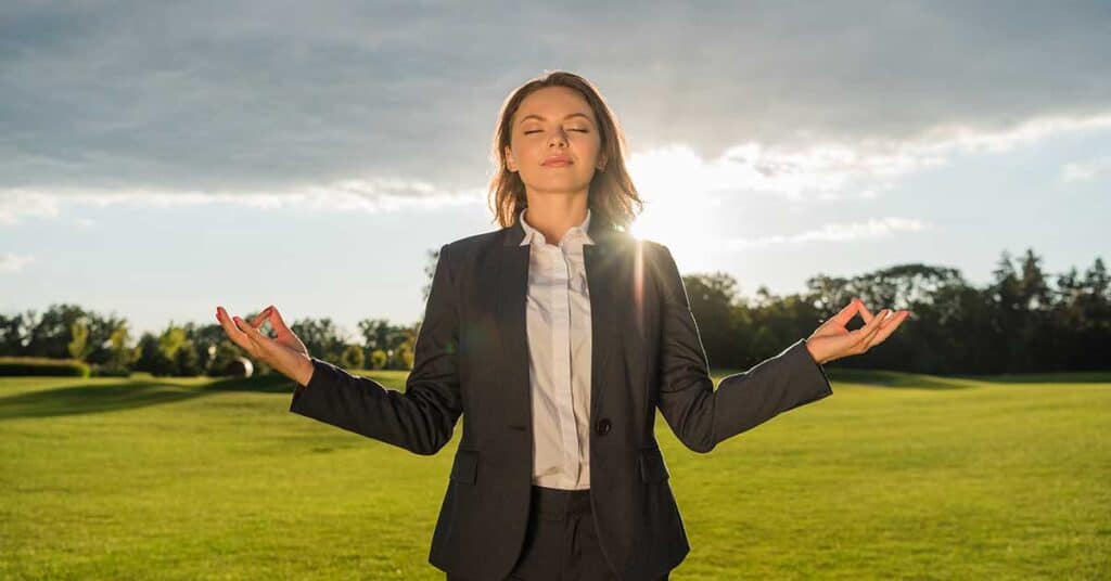 A woman in a business suit meditates outdoors on grass, eyes closed, drawing in the sun’s warm energy as it shines in the background.