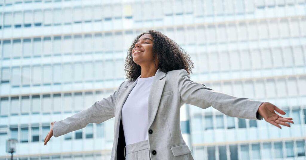 A woman in a light gray suit stands outside with her arms outstretched and eyes closed in front of a modern glass building, embracing a moment to reset.