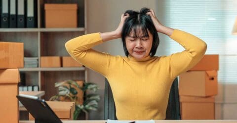 An executive woman in a yellow sweater sits at a desk with her hands on her head, eyes closed, appearing frustrated or stressed. Cardboard boxes and office supplies are scattered around her.