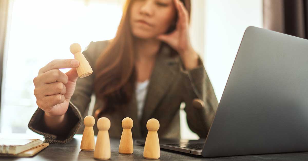 A woman sits at a desk holding a wooden figure in one hand, with other figures lined up in front of an open laptop, thoughtfully exploring career possibilities.