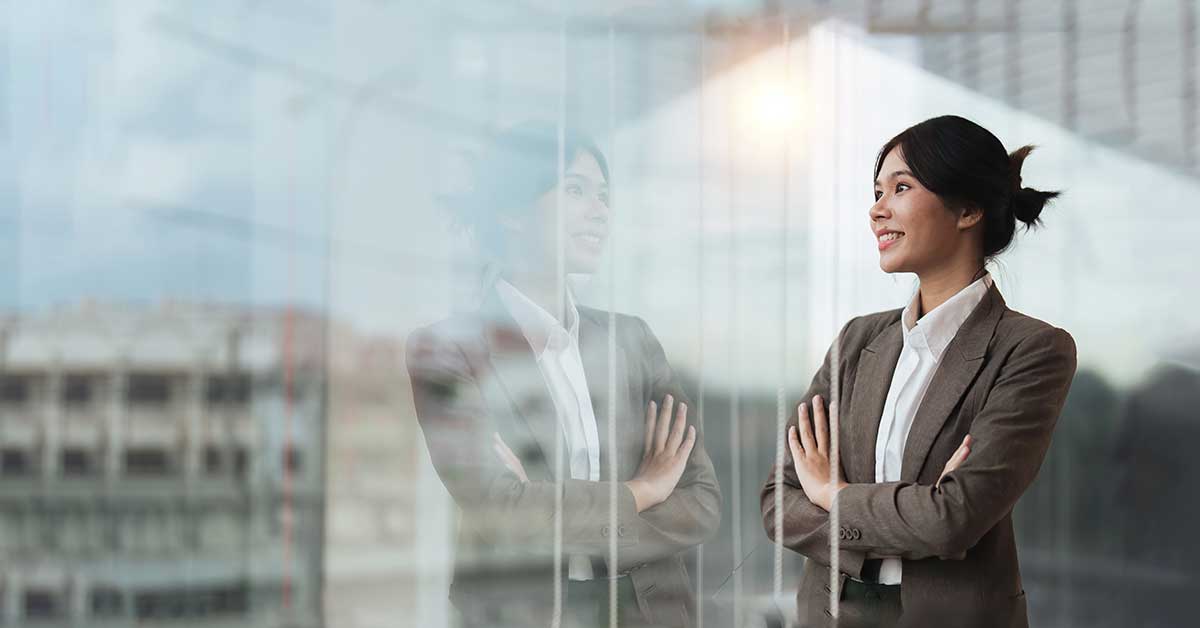 A woman in a suit stands with arms crossed, smiling, looking out a window; her reflection on the glass highlights her visibility and confidence.