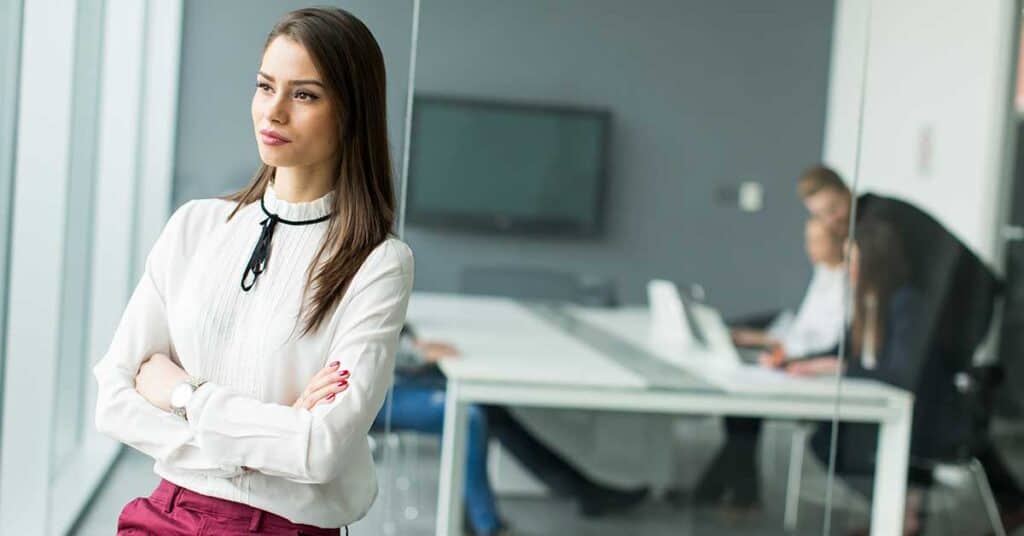 A woman stands with folded arms near a glass wall, exuding confidence and alignment, while three people sit at a conference table in the background of a modern office.
