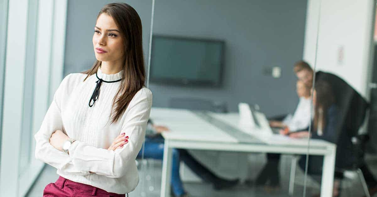 A woman stands with folded arms near a glass wall, exuding confidence and alignment, while three people sit at a conference table in the background of a modern office.