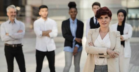 A woman in business attire stands confidently with arms crossed, exemplifying leadership as she leads a diverse group of five colleagues outdoors.