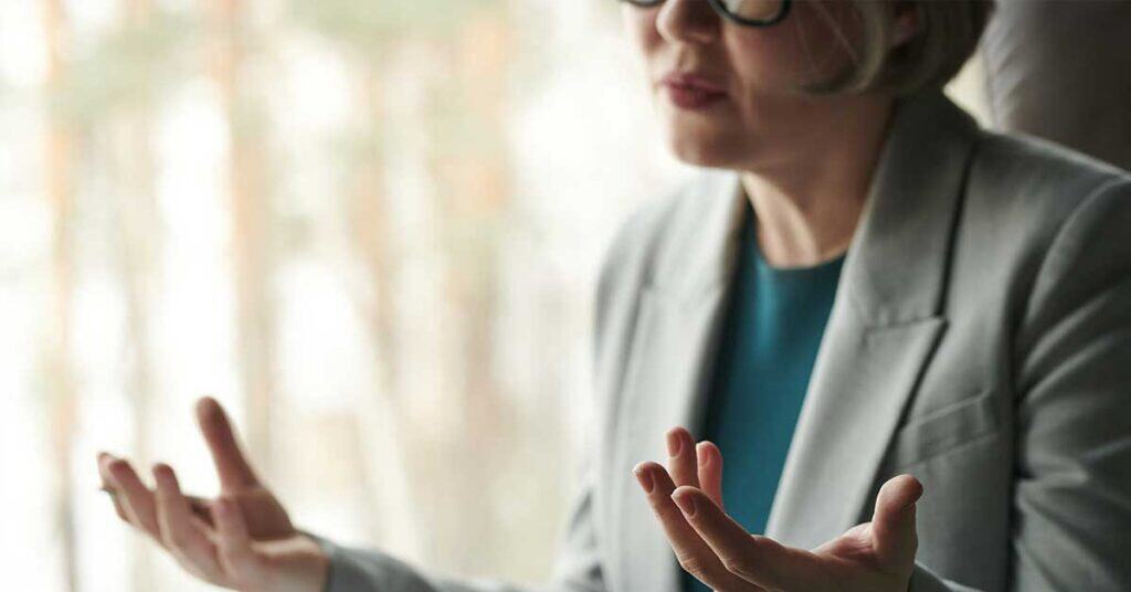 A person in a gray blazer and glasses gestures with open hands, embodying leadership while seated indoors against a blurred background.