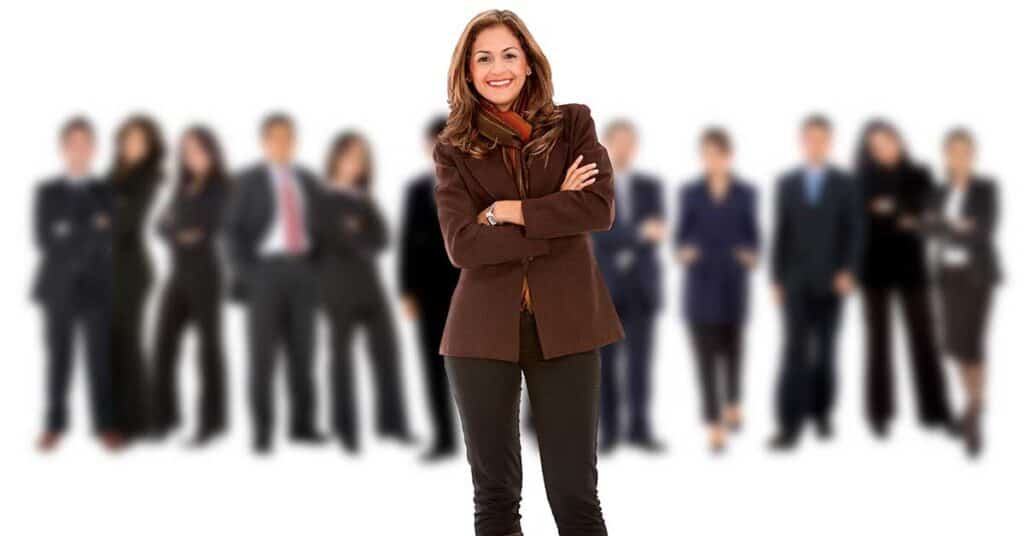A woman in business attire stands confidently with arms crossed, showcasing her influence in front of a blurred group of people in professional clothing.