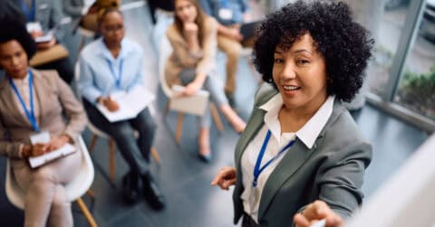A woman in a suit stands at the front of a room, demonstrating leadership as she speaks to a seated audience who are taking notes and listening attentively.