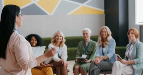 A woman holding papers addresses a group of five seated women, who are listening attentively in a brightly lit room during a mentoring session.