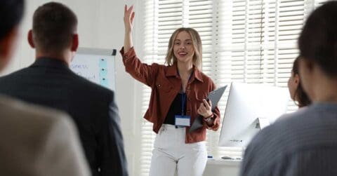 A woman demonstrates leadership as she stands and gestures while presenting to a small group in an office setting with a whiteboard and computer visible.