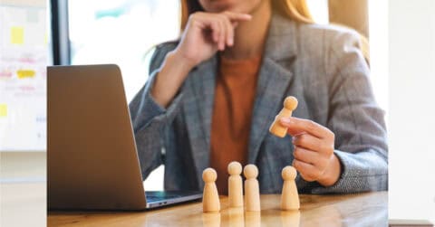 A person in a blazer sits at a desk with a laptop, holding a wooden figure among several others, highlighting the concept of leadership and decisive decision-making.
