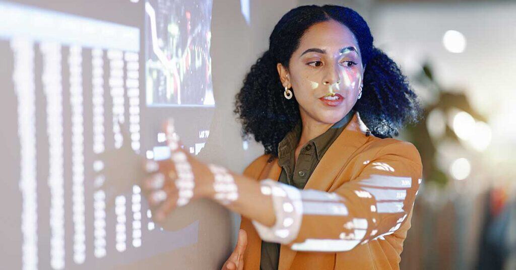 A woman in a brown blazer points at AI-driven data projected onto a wall, with charts and graphs visible on the projection.