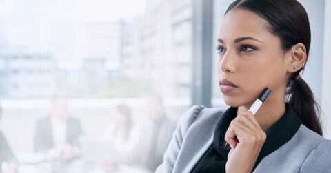 A woman in business attire holds a marker to her chin and looks thoughtful, contemplating ways to boost speed, with a blurred office meeting in the background.