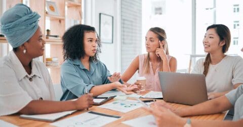Five professional women sit around a table with papers, charts, and a laptop, engaged in a dynamic business discussion in a bright office setting.