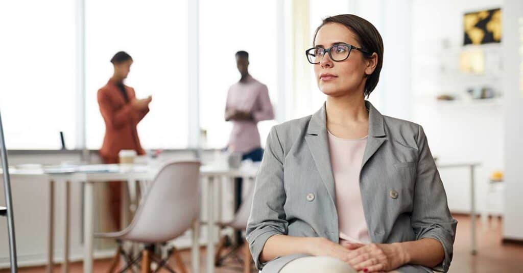 The Quiet Loneliness Of Being The One Everyone Counts On A woman wearing glasses sits in an office chair, looking to the side, with two people standing and talking in the background.