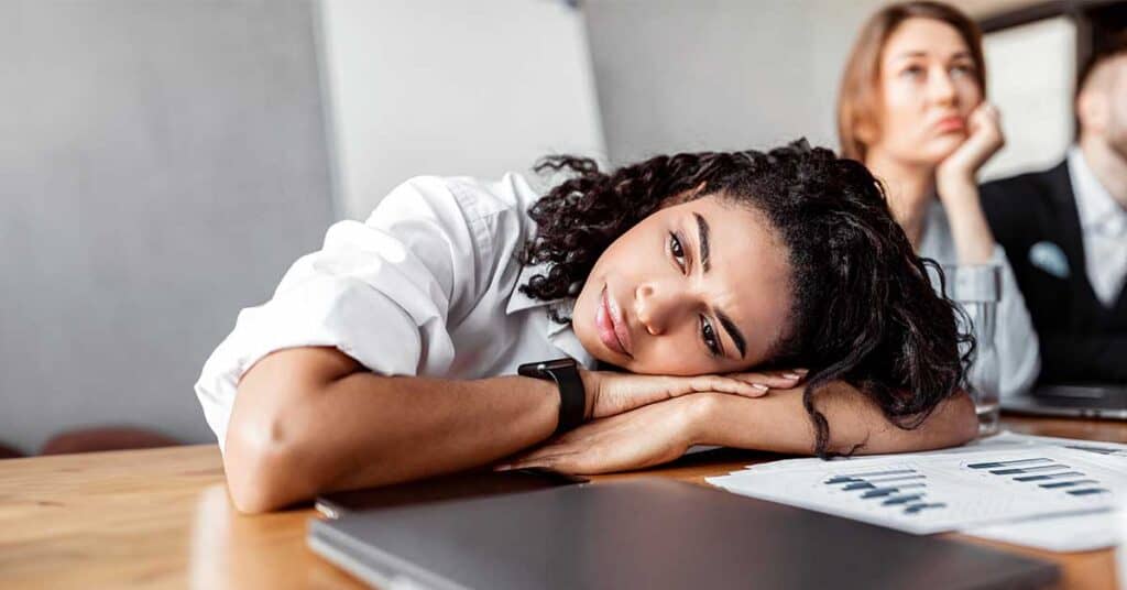 The Exhaustion Of Being Easy To Work With A woman rests her head on her arms at a desk during a meeting, appearing tired, with charts and a laptop in front of her.