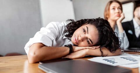A woman rests her head on her arms at a desk during a meeting, appearing tired, with charts and a laptop in front of her.
