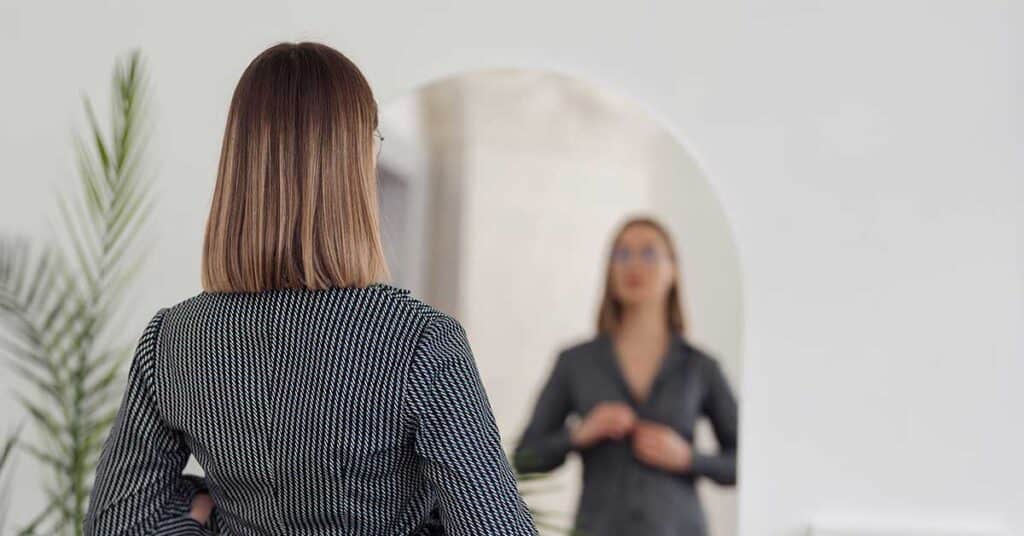 The Pressure To Be The Same Person Forever A woman with shoulder-length hair stands in front of a mirror, adjusting her suit jacket.