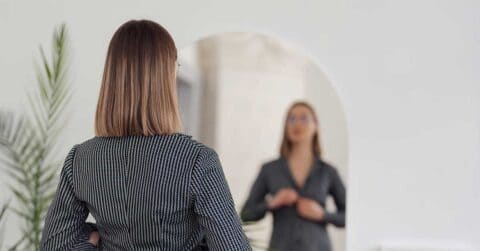 A woman with shoulder-length hair stands in front of a mirror, adjusting her suit jacket.